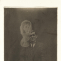 Black and white phpotograph of a seated man showing a spectral female head above him