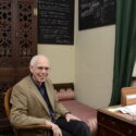 Colour photograph of Jim Bennett sat at desk with blackboards hung behind
