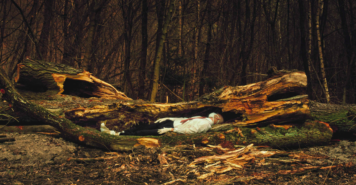 Image of a fragmented fallen tree, in which a man in a white shirt and black trousers lies as though asleep or unconscious. He wears a surgical mask. In the background is a dark forest.